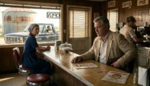 A Diner Counter, 1980s-Style, Photorealistic Cinematic Style, Wide-Angle Shot. Foreground: A sturdy, middle-aged Austrian man (50s) with short graying hair, wearing a slightly rumpled 1980s summer sports jacket over an open-collared shirt, sits on a worn red faux-leather bar stool. He stares blankly at the polished, sticky formica counter with a stoic, melancholic expression. One hand rests on the counter, while a bulky leather pouch bulges subtly inside his jacket pocket. Next to him on the counter sits a laminated Diner menu card featuring a retro cartoon mascot with unnaturally bright, glowing, aggressive white teeth (Zahnweiß). Midground: Two stools down, a woman in her late 40s (Brenda) with a weather-worn, serene face, wearing a plain, modest dark blue cotton dress and a faded headscarf (Pennsylvania-Dutch style), quietly turns her head to look at him with a calm, observant gaze. Behind the counter, a waitress in a pale uniform works in a soft blur. Background: Through the grease-stained, sun-lit diner window, a black Ford pickup truck is parked in the glaring Indiana midday heat near a rusted grain silo. Inside the truck, a shadowy male figure wearing a camouflage cap stares intensely back toward the diner counter. Lighting & Mood: Warm, dusty afternoon sunlight filtering through Venetian blinds and dirty glass, casting long shadows and highlights on dust motes in the air. Gritty, cinematic 35mm film grain, muted color palette of faded rust-reds, mustard yellow, and warm wood tones, evoking a grounded 1980s crime-drama aesthetic.
