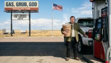 Ein älterer, sichtlich überforderter Mann in traditioneller österreichischer Trachtenjacke steht an einer entlegenen Tankstelle im flachen, unendlichen Nirgendwo von Nebraska. Neben einem riesigen weißen Ford-Pickup hält er ungläubig die Zapfpistole in der einen Hand und schaut mit entsetztem Blick direkt in die Kamera. Unter dem armseligen Schatten des Tankstellendachs umklammert er wie einen Eimer einen gigantischen, dampfenden Pappbecher mit der Aufschrift „Triple-Shot HUGE American Java“. Im Hintergrund erstreckt sich eine schnurgerade Landstraße bis zum Horizont, überragt von einer mächtigen US-Flagge und einem verblichenen Reklameschild mit der Aufschrift „GAS, GRUB, GOD – LAST CHANCE FOR CARAMEL LATTE“.