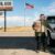 Ein älterer, sichtlich überforderter Mann in traditioneller österreichischer Trachtenjacke steht an einer entlegenen Tankstelle im flachen, unendlichen Nirgendwo von Nebraska. Neben einem riesigen weißen Ford-Pickup hält er ungläubig die Zapfpistole in der einen Hand und schaut mit entsetztem Blick direkt in die Kamera. Unter dem armseligen Schatten des Tankstellendachs umklammert er wie einen Eimer einen gigantischen, dampfenden Pappbecher mit der Aufschrift „Triple-Shot HUGE American Java“. Im Hintergrund erstreckt sich eine schnurgerade Landstraße bis zum Horizont, überragt von einer mächtigen US-Flagge und einem verblichenen Reklameschild mit der Aufschrift „GAS, GRUB, GOD – LAST CHANCE FOR CARAMEL LATTE“.